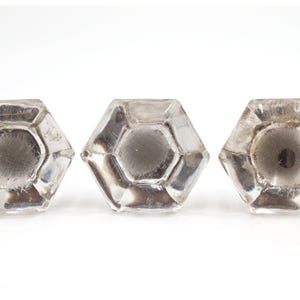 Puede incluir: Tres pomos de cristal transparente y facetado con forma hexagonal. Cada pomo tiene un centro plateado y está colocado sobre un fondo blanco. Los pomos son probablemente para muebles o uso decorativo.