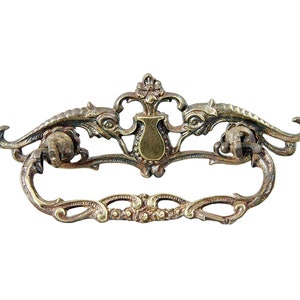 Puede incluir: Tirador de cajón de latón adornado con un diseño decorativo que presenta dos dragones estilizados y un escudo central. El tirador tiene una manija lisa y curva.