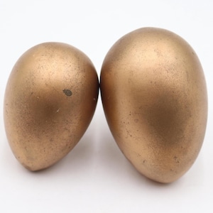 May include: Two bronze-coloured, egg-shaped decorative objects. The objects have a textured surface and a metallic sheen. They are displayed against a white background. The eggs are similar in size and shape.