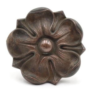 Könnte beinhalten: Ein dunkelbrauner, metallischer Möbelknopf in Form einer fünfblättrigen Blume. Die Oberfläche hat ein verwittertes, mattes Finish mit einer zentralen kreisförmigen Erhebung und erhabenen, abgerundeten Blütenblättern, die vom Mittelpunkt ausstrahlen.