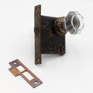 May include: An antique door knob set with a clear, faceted glass knob and a dark, rusty metal backplate. A separate, matching metal strike plate is also present. The hardware is likely vintage and shows signs of wear.