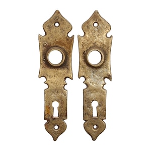 Puede incluir: Dos escudos de pomo de puerta de latón antiguo con diseños ornamentados y agujeros para llaves.