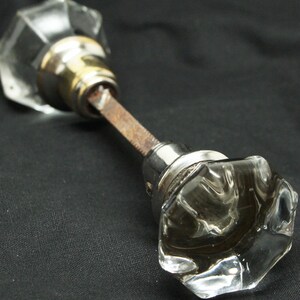 May include: Clear glass door knob with a faceted design. The knob has a metal base with a tarnished, rusty rod connecting the two knobs. The hardware is set against a dark background.