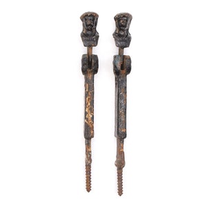May include: Two antique black cast iron gate hinges with a decorative human head design. The hinges are rusty and have a screw at the bottom.