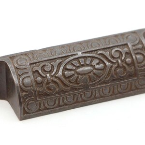 May include: A vintage cast iron drawer pull with an ornate design. The pull is a dark brown color and has a slightly curved shape.