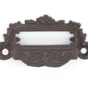 May include: Ornate, cast iron drawer pull with a rectangular label holder. The dark brown pull features a decorative floral and leaf design. The pull has two holes for mounting and is set against a white background.