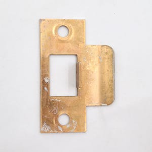 May include: A rectangular, brass-colored door strike plate with a rectangular opening and two circular holes. The plate has a bent, right-angled extension. The metal shows signs of wear and age.