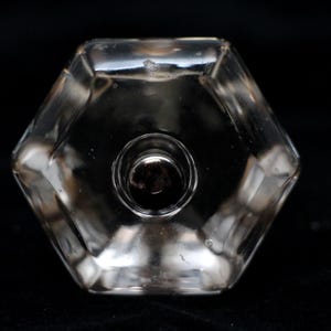 Pode incluir: Um tampão de garrafa de vidro transparente com forma hexagonal e uma abertura redonda no centro. O tampão tem uma superfície lisa e polida.