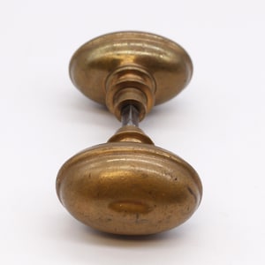 May include: Antique brass door knob set with an oval shape. The door knob set has a tarnished finish and a connecting rod. The door knobs are approximately 3 inches long.