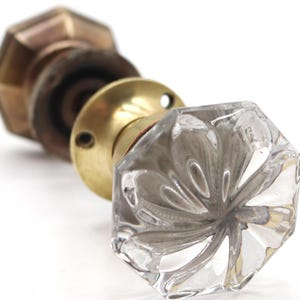 Könnte beinhalten: Nahaufnahme eines Vintage-Türknaufs mit einem klaren, facettierten Glasgriff. Der Knauf hat einen Messingfuß und eine sechseckige Form. Das andere Ende des Knaufs ist ein dunkelbraunes Metall mit einer sechseckigen Messingform.