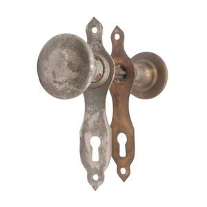 May include: Two antique door knobs with keyhole plates. One knob is a tarnished silver color, and the other is a brass color. The door knobs have a vintage, weathered appearance, suggesting age and use. The keyhole plates have decorative shapes.