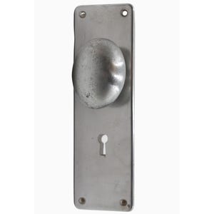 Puede incluir: Un pomo de puerta plateado con una manija redonda ligeramente abovedada y una cerradura. El pomo está unido a una placa rectangular.