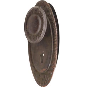 May include: Antique bronze door knob and backplate. The oval backplate and round knob feature ornate detailing. The knob has a circular design around the center. The keyhole is centered on the backplate. The hardware has a weathered appearance.