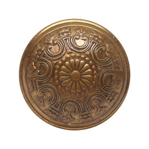 May include: A round, ornate, brass button with a raised, floral design. The button has a circular, embossed pattern with a central flower motif.