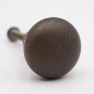 May include: A close-up of a bronze-colored, round cabinet knob with a short, cylindrical stem. The knob has a smooth, matte finish and is set against a white background. This hardware is suitable for drawers and cabinets.