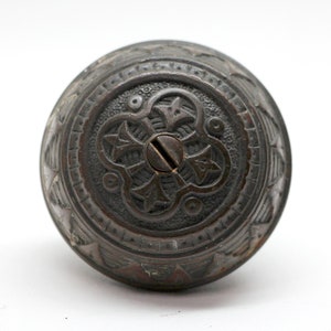 May include: A vintage, ornate, dark bronze door knob with a decorative, intricate design. The knob has a keyhole and a screw in the centre.