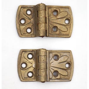 May include: Two antique brass hinges with a decorative floral design. Each hinge has a rectangular plate with rounded corners and three circular holes. The central barrel is textured. The hinges are likely for a cabinet or small door.