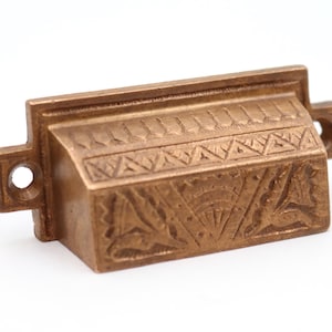 Może przedstawiać: Mosiężny uchwyt do szuflady z misternymi, wytrawionymi wzorami. Prostokątny uchwyt ma szczegółowy wzór z przodu i po bokach, z małymi otworami z boku do montażu. Okucia są prawdopodobnie przeznaczone do szafki lub szuflady.