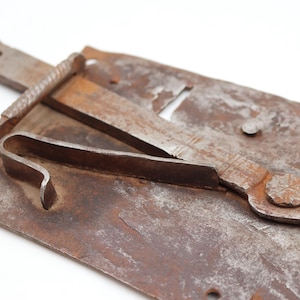 May include: Close-up of a rusty, antique metal latch. The latch features a rectangular base with a curved lever and a sliding bolt. The metal has a weathered, aged appearance with visible rust and signs of wear, suggesting it is an old piece.