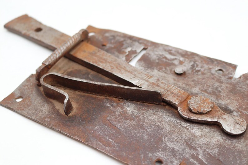 May include: Close-up of a rusty, antique metal latch. The latch features a rectangular base with a curved lever and a sliding bolt. The metal has a weathered, aged appearance with visible rust and signs of wear, suggesting it is an old piece.