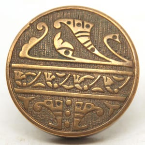 Puede incluir: Un botón redondo de color bronce con un diseño en relieve. El diseño presenta formas y patrones estilizados, incluyendo líneas curvas y elementos geométricos. El botón tiene una superficie texturizada y una estética vintage.