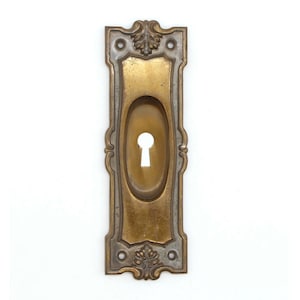 May include: Antique brass keyhole cover with an ornate design and a keyhole in the center.