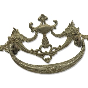 Può includere: Una maniglia per cassetto antica in ottone decorata con un motivo a volute. La maniglia ha una forma curva e un ovale centrale.