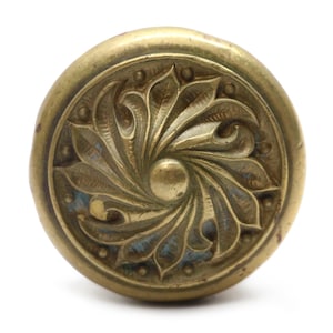 Könnte beinhalten: Antiker Schrankknauf aus Messing mit einem verzierten floralen Design. Der Knauf hat ein zentrales kreisförmiges Element, das von stilisierten Blättern und kleinen Perlenakzenten umgeben ist. Das Metall hat eine leicht gealterte Patina.