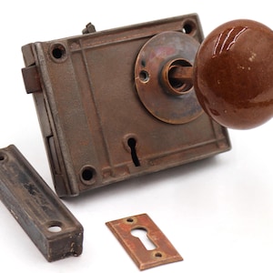 May include: An antique door lock set with a brown ceramic doorknob. The set includes a rectangular lock body, a strike plate, and a keyhole cover, all with a weathered, rusty appearance. The hardware is likely made of metal.