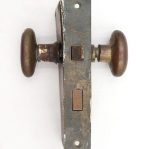 May include: An antique door lock set with oval bronze knobs and a rectangular, aged metal plate. The lock mechanism is visible, showing a rectangular bolt and strike plate. The hardware has a weathered appearance, suggesting age and use.