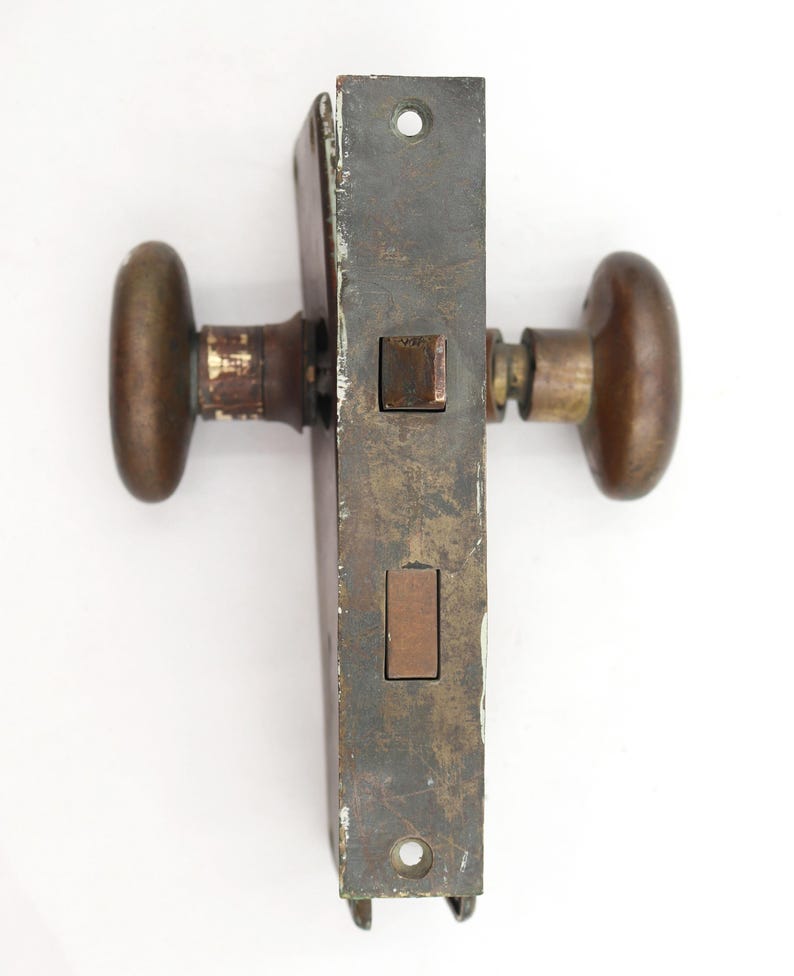 May include: An antique door lock set with oval bronze knobs and a rectangular, aged metal plate. The lock mechanism is visible, showing a rectangular bolt and strike plate. The hardware has a weathered appearance, suggesting age and use.