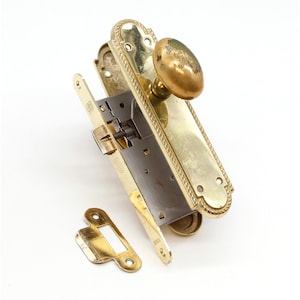 May include: A brass door knob set with a round knob and a rectangular plate with a rope design. The plate has a keyhole and a small latch. The set is in a disassembled state.