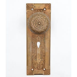 Puede incluir: Una placa de puerta rectangular de latón con un pomo redondo a juego. La placa presenta un intrincado patrón floral en relieve y una cerradura central. El pomo tiene un diseño circular de rayos de sol con grabados detallados en la superficie.