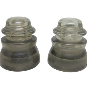 May include: Two vintage glass insulators with a gray tint. The insulators have a rounded top and a wide base. The base of each insulator has the text "NEW YORK INSULATOR CO.  U.S.A." printed on it.