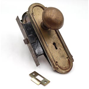 Peut inclure: Ensemble de quincaillerie de porte ancienne comprenant un bouton de porte et une plaque arrière en laiton, une serrure à mortaise en métal noir et une gâche séparée. Le bouton de porte est rond, de couleur bronze vieilli. Probablement du début du XXe siècle.