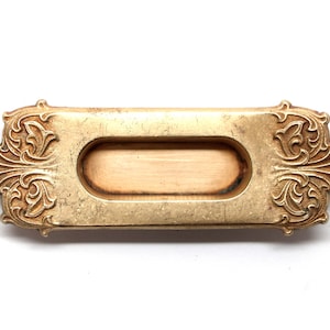 Puede incluir: Un tirador de puerta de metal dorado con un diseño ornamentado. El tirador tiene una forma rectangular con una abertura ovalada hundida.