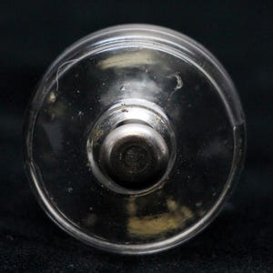 Könnte beinhalten: Ein durchsichtiger Glasflaschenverschluss mit einem silbernen Metalldeckel. Der Verschluss ist rund und hat eine glatte Oberfläche.