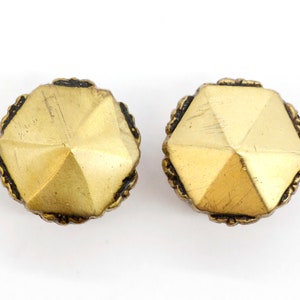 May include: A pair of gold-toned metal earrings with a geometric, faceted design. The earrings have a black border around the edges.