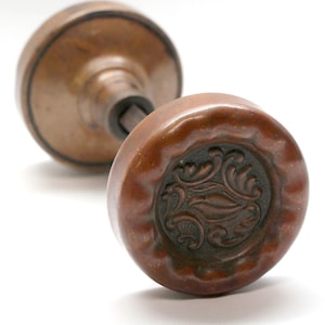 Puede incluir: Dos pomos de puerta antiguos de color marrón con un diseño floral decorativo. Los pomos están hechos de un material marrón oscuro y tienen un patrón detallado en relieve en el centro. Los pomos están sobre un fondo blanco.