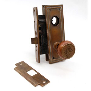 May include: Antique brass door lock set with a wooden knob featuring a diamond-shaped logo. The lock has a keyhole and a latch. The knob is attached to a square plate with a decorative border.