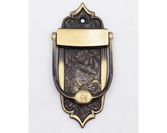 May include: Ornate bronze door knocker with a detailed, textured design. The knocker features a rectangular plate above a curved handle and a decorative base. The metal has a dark, aged appearance with gold accents.