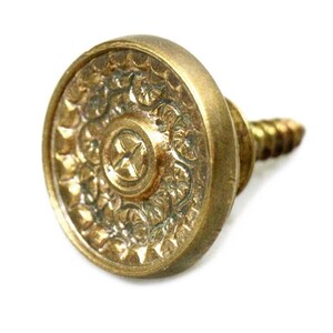 Puede incluir: Un pomo de metal dorado con un diseño floral decorativo y un tornillo. El pomo es redondo y tiene un centro elevado.