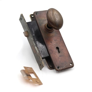 May include: An antique door lock set with a tarnished bronze finish. The set includes a rectangular plate with a keyhole, an oval doorknob, and a separate strike plate. The hardware is likely vintage.