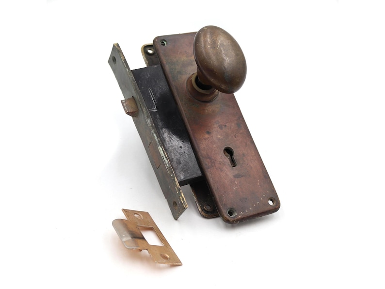 May include: An antique door lock set with a tarnished bronze finish. The set includes a rectangular plate with a keyhole, an oval doorknob, and a separate strike plate. The hardware is likely vintage.