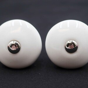 May include: Two white ceramic cabinet knobs with a glossy finish. Each knob features a silver-colored, circular metal accent in the center. The knobs are round and smooth, suitable for furniture or decorative projects.