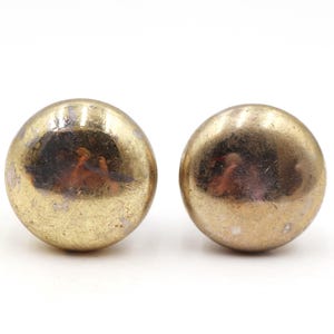 May include: Two antique brass knobs with a tarnished, aged appearance. The knobs are spherical and have a warm, golden hue with darker spots, suggesting age and use. They are set against a plain white background.