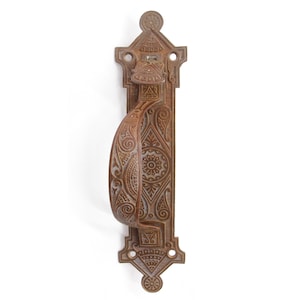 Op de afbeelding: Een sierlijk, antiek deurbeslag met een decoratief, ingewikkeld ontwerp. De hendel is gemaakt van metaal en heeft een bruine patina-afwerking.