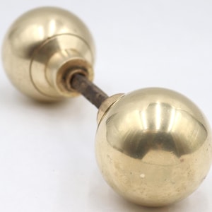Puede incluir: Un juego de manijas de puerta de latón antiguo con dos perillas esféricas conectadas por una varilla de metal oscuro. Las perillas tienen una superficie pulida y reflectante, con un diseño simple y clásico.