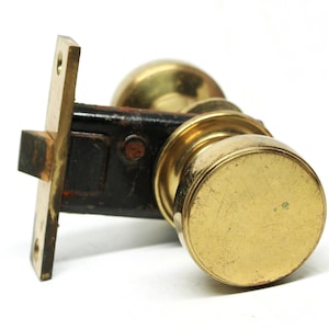 Könnte beinhalten: Ein Messing-Türknopf mit einem schwarzen Riegel. Der Knopf ist rund und hat eine glatte Oberfläche. Der Riegel ist rechteckig und hat ein kleines Loch in der Mitte.