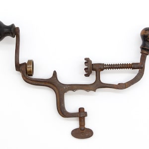 Puede incluir: Una herramienta de metal oxidado con dos mangos de madera y un mecanismo de resorte. La herramienta se utiliza para apretar o aflojar objetos.